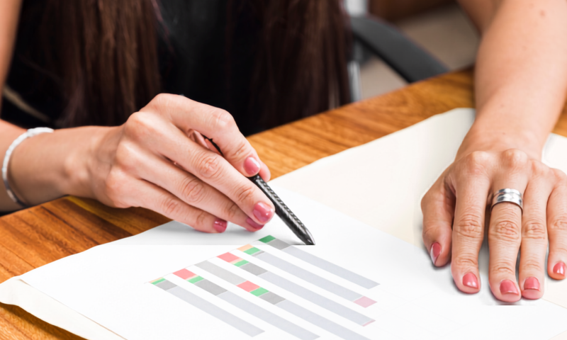 Close-up view of a person seated at a wooden desk, holding a black pen and pointing to a horizontal bar chart on a printed document. Their other hand rests beside the paper on the table.