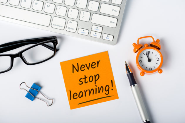 Photograph of keyboard, glasses, paper clip, post-it note (which reads: "never stop learning"), pen, and clock.