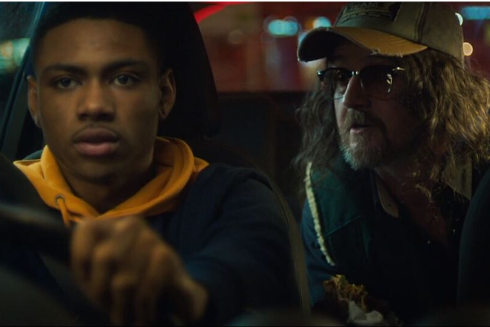Black man sat in a car with another man staring at him behind.