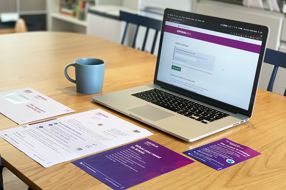 A laptop showing the Census 2021 questionnaire start page and a collection of census materials including an initial contact letter with the access code, a ‘What you need to know’ flyer, a Census 2021 branded envelope and Census postcard. All are displayed on a kitchen table.
