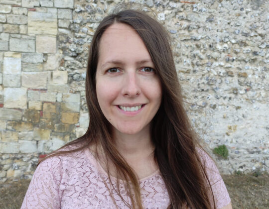 Rebecca Jakeway, woman smiling with long brown hair and a pink t-shirt, in front of a stone wall outside.