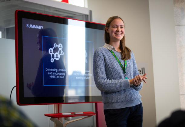 Vera Pudilova Communications and Engagement Adviser, is standing in the centre of a room presenting to an audience with a screen behind her.