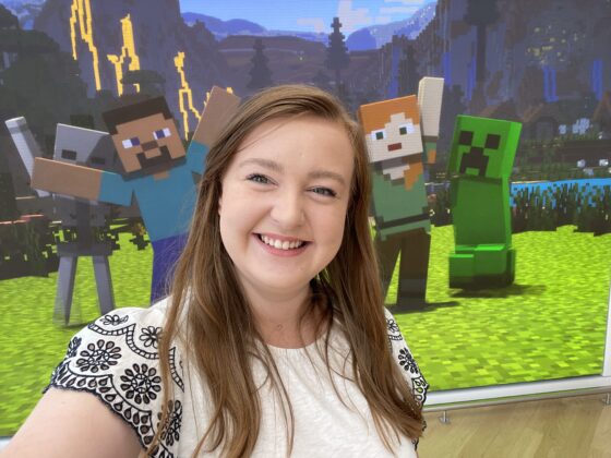 A selfie image of Abby Wade, GCS Applied Innovation Manager, at the Microsoft Experience Centre in London. Abby is smiling at the camera and there is a scene of Minecraft characters in the background.