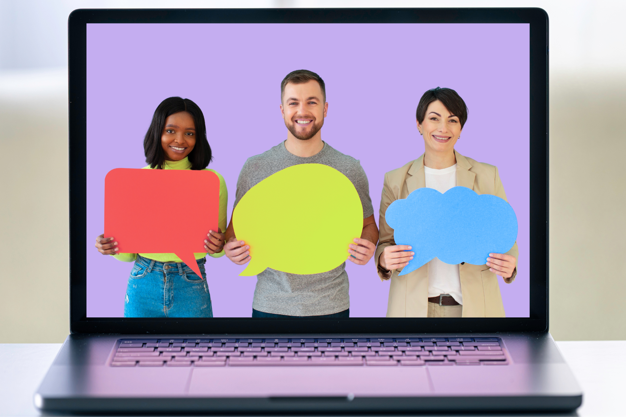 A laptop screen with a light purple background displaying three people standing side by side and smiling at the camera while holding colourful speech bubble cutouts: a red rectangle, a yellow oval, and a blue cloud shape.