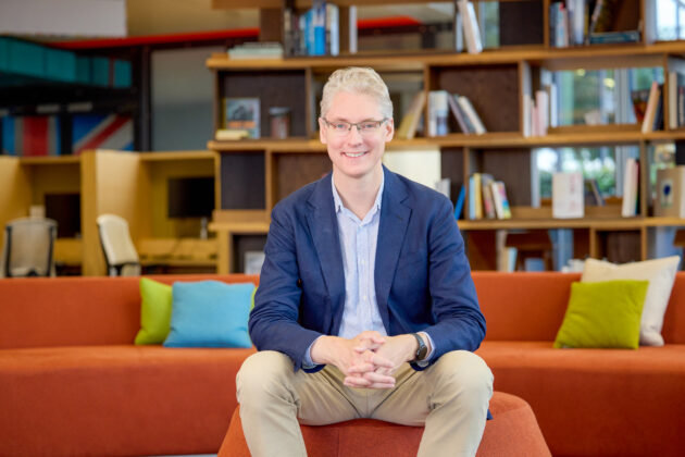 David Black sitting on a stool smiling towards the camera. There is a seating area, book shelves and desks in the background