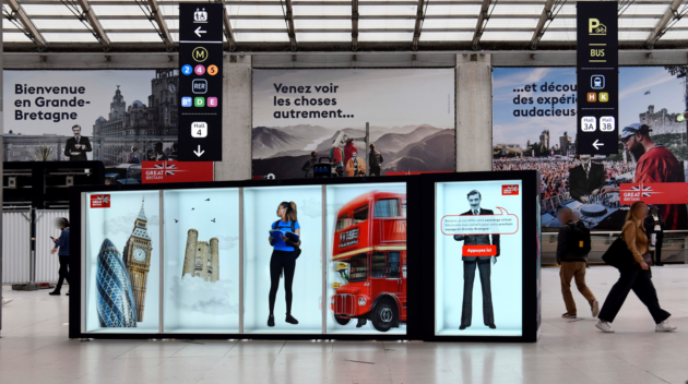 A busy indoor station with large promotional signs showcasing British tourism. In the background, three large posters with French-language text read: “Bienvenue en Grande-Bretagne”, “Venez voir les choses autrement…” and “...et découvrez des experiences audacieuses”. Which translates to: “Welcome to Great Britain. Come see things differently… and discover daring experiences”. In the foreground a digital display shows images associated with the UK such as the Gherkin, Big Ben and a red double decker bus. Overhead signage shows transport icons for metro lines, buses and parking while several people walk through the station.