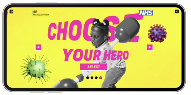 A vibrant NHS and HM Government poster designed in the style of a mobile video game character selection screen. The background is bright yellow with large magenta text reading “CHOOSE YOUR HERO” and a “SELECT” button below. At the centre is a black and white photo of a young girl holding a toy pugil stick, as if preparing for battle.  Illustrations resembling flu particles appear on either side of her. There are navigation arrows and a row of dots indicating multiple selection screens.