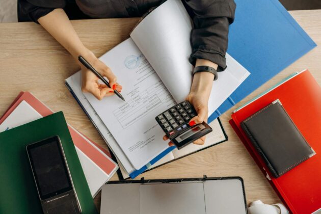 A person is working at a desk with open documents, holding a calculator, and writing with a pen. Folders, a laptop, and a notebook surround them.