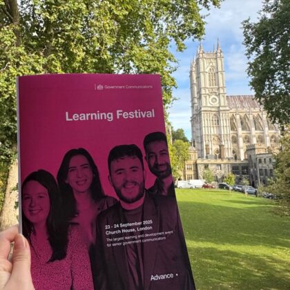 A person holding a bright pink programme in front of green space and a cathedral in the background. The programme reads:Learning Festival. 23 to 24 September 2025. Church House, London. The largest learning and development event for senior government communications.
There is a a photo of four people on the cover. The Government Communications logo is at the top and the Advance logo at the bottom of the programme.