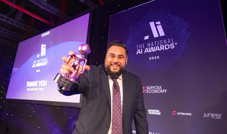 A person holds a purple robot award on a stage. The backdrop is purple with branding from 'The National AI Awards'.