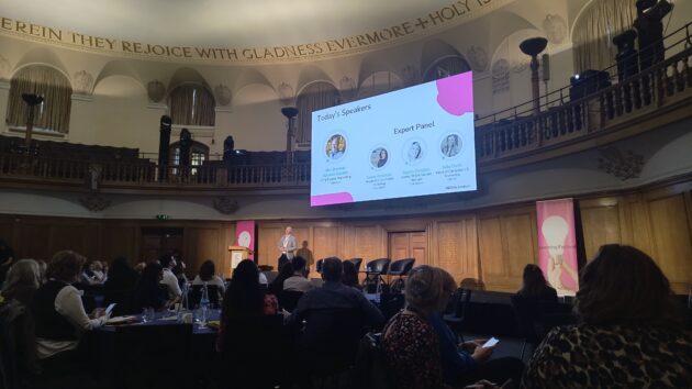 A stage with a man talking and a room of people. A large screen shows four people and the words Today's Speakers.