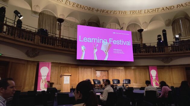 A stage and with a large screen on the wall displaying a pink slide and the words Learning Festival on it.