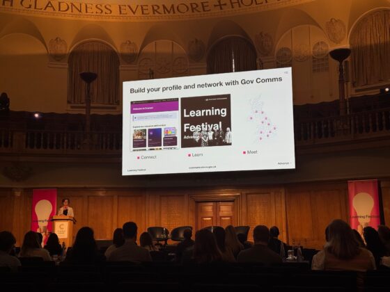 Image showing the stage with people in front of it and a large screen showing: Build your profile and network with Gov Comms. Connect. Learn. Meet