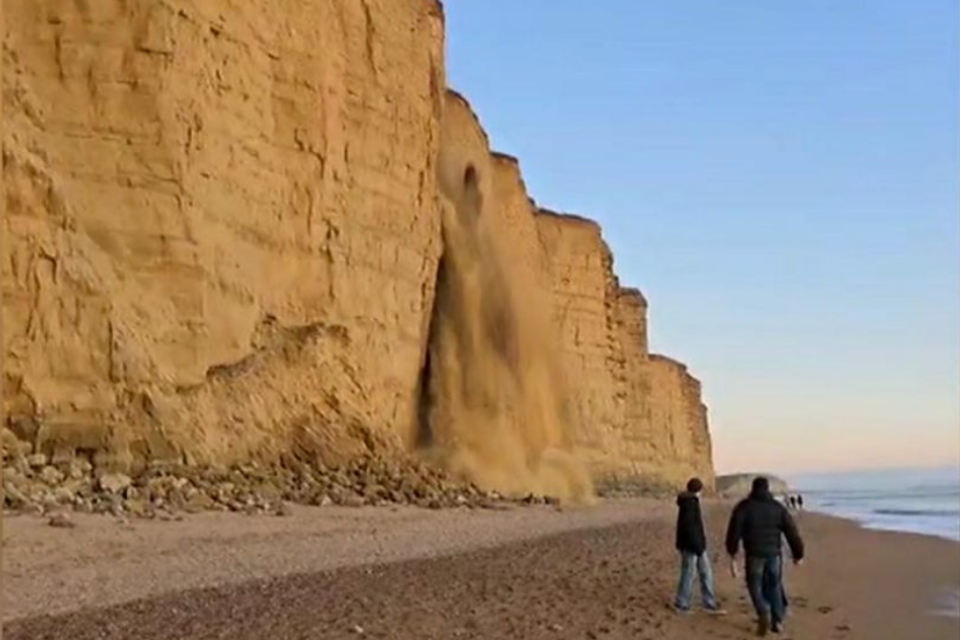 A dramatic photograph of the unexpected rockfall on the Jurassic Coast, the event which inspired this blog post on viral communication. In the foreground there are two people on the beach, and there is blue skies, the sea and more people walking along the beach in the background.