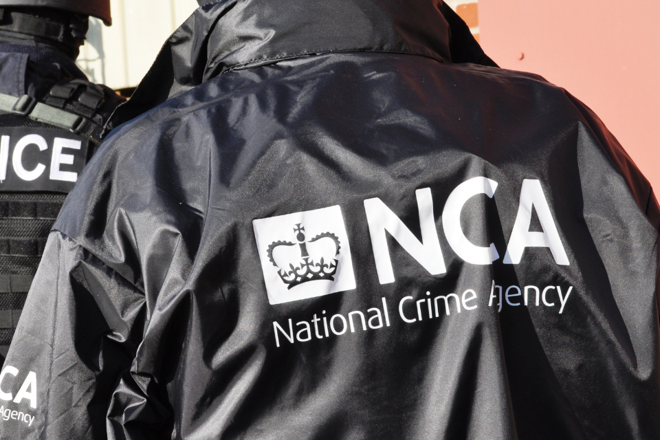 Two people with their backs towards the camera. Their uniformed jackets have text in white, one with “Police” and the other with “NCA, National Crime Agency”.