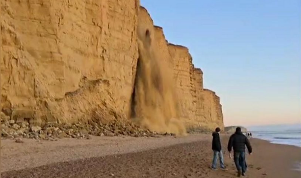 A dramatic photograph of the unexpected rockfall on the Jurassic Coast, the event which inspired this blog post on viral communication. In the foreground there are two people on the beach, and there is blue skies, the sea and more people walking along the beach in the background.
