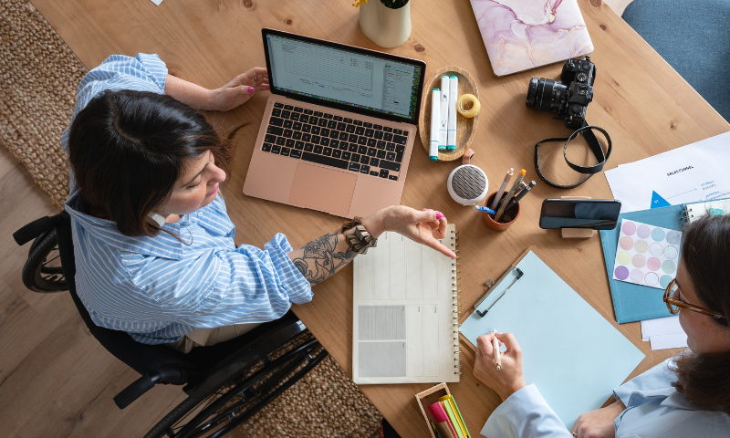 A person in a wheelchair at a desk that has a laptop, a camera and papers on it. There is a person beside them writing on a clipboard.