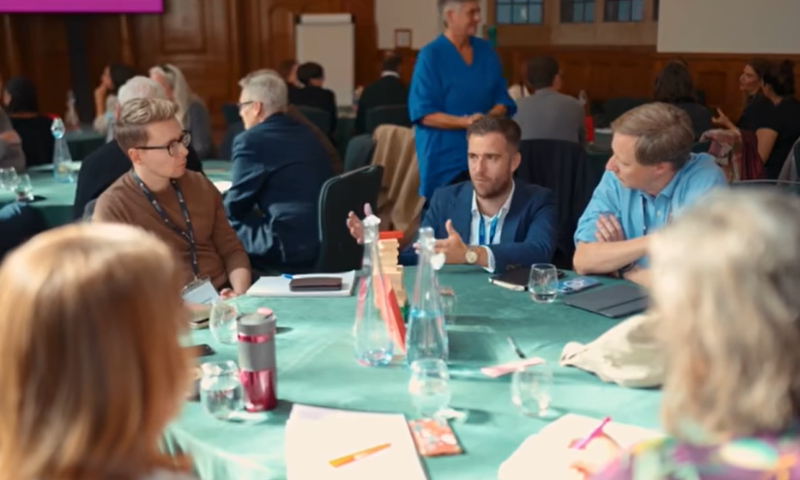 A group of five people sit around a large table which has papers and pens on it. They are listening to one member of the group speaking. Behind them are more groups of people working at tables.