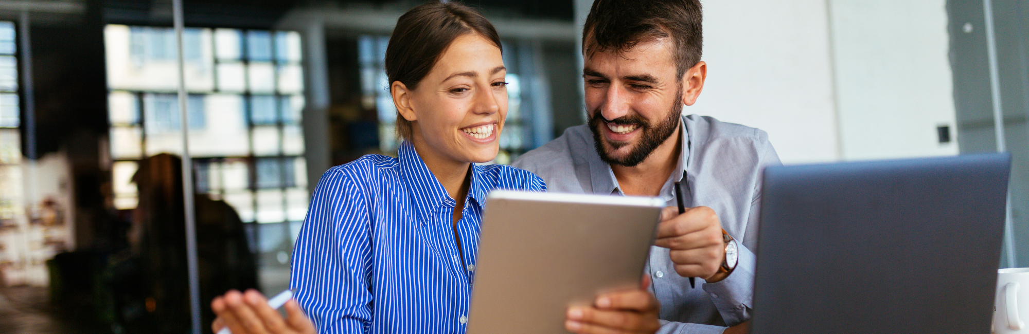 Two people sit side by side in front of a laptop. One person is holding a tablet and they both look at the screen.