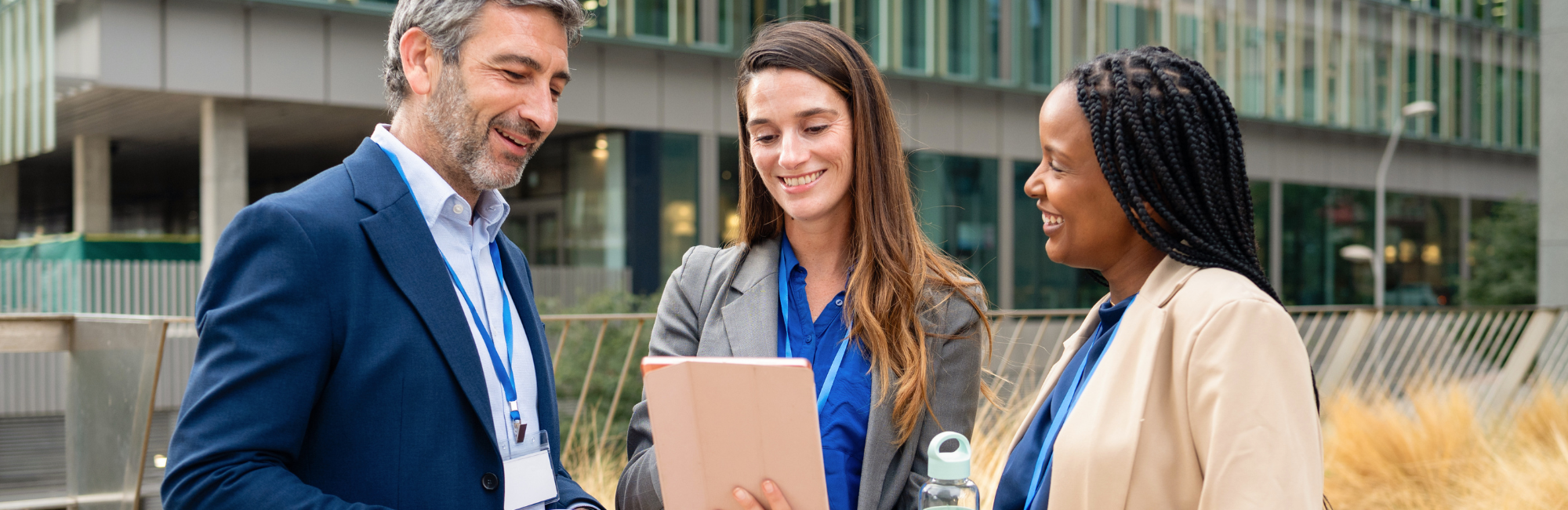 Three people standing together in front of a building and smiling while looking at a tablet.
