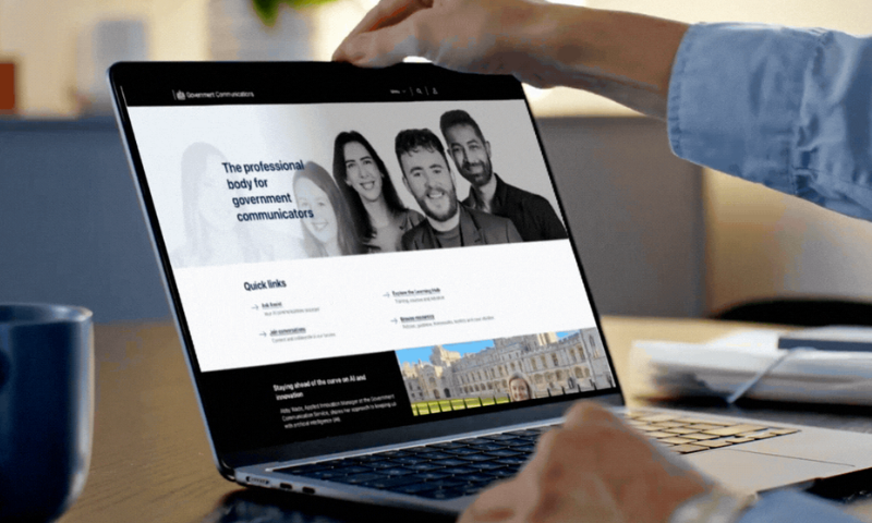 A person is opening a laptop on a desk. The laptop screen displays the Government Communications website. Across the top of the page is a landscape photo of five people. Beneath it is a 'Quick links' section