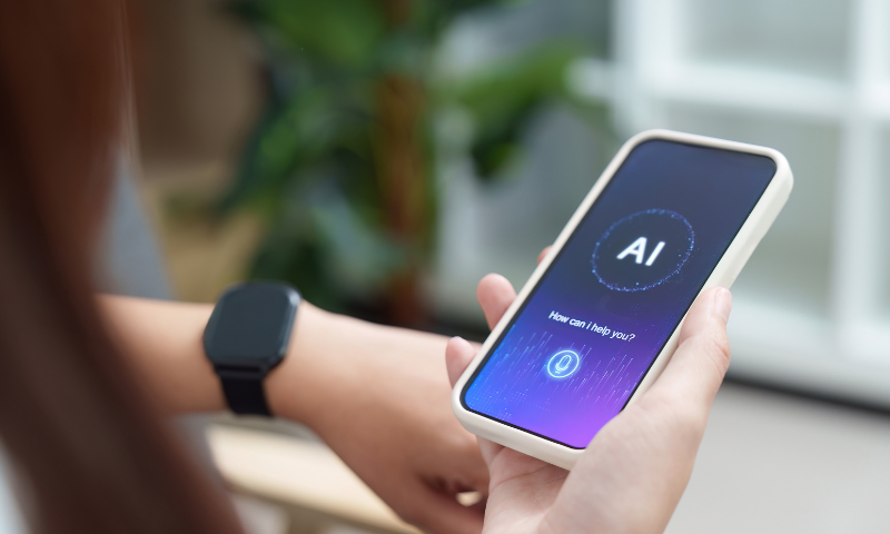 A person holds a mobile phone which displays an AI with text: AI, How can I help you? The screen is a gradient purple with a blue button of a microphone at the bottom.