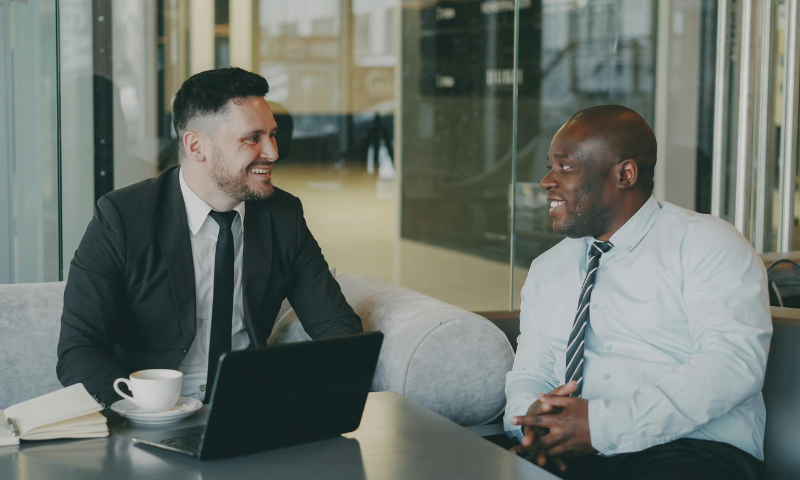 Two people sitting at a table together where there is a laptop and a cup of coffee on it.